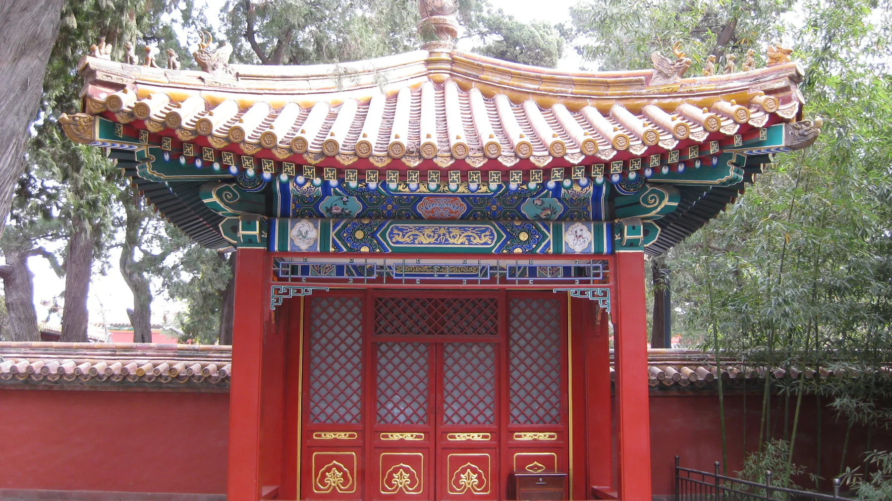 An ornate red gateway with closed lattice doors and an upturned tiled roof stands against a red wall beneath trees.