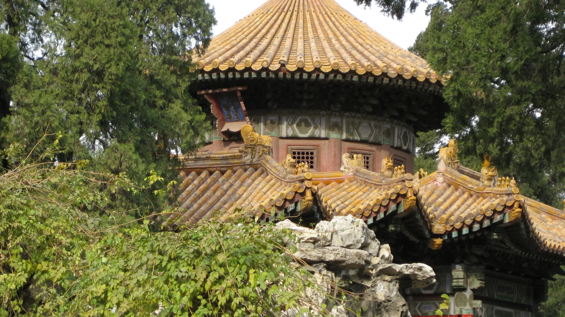 A pavilion with a rounded golden roof rises above smaller tiled roofs, rocks and dense green planting.