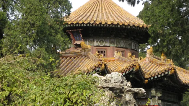 A pavilion with a rounded golden roof rises above smaller tiled roofs, rocks and dense green planting.