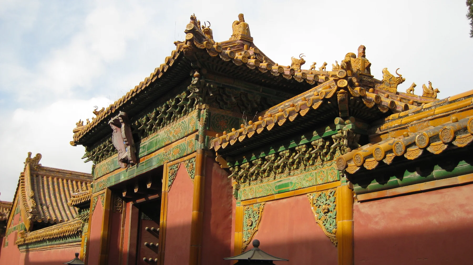 An ornate gateway and red wall are topped with golden tiles, curved roof corners and green decorative panels.