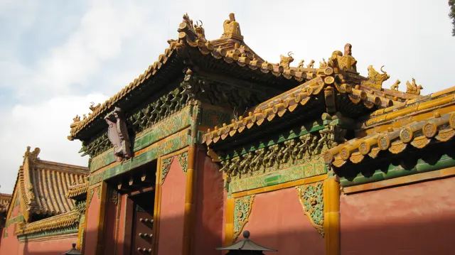 An ornate gateway and red wall are topped with golden tiles, curved roof corners and green decorative panels.