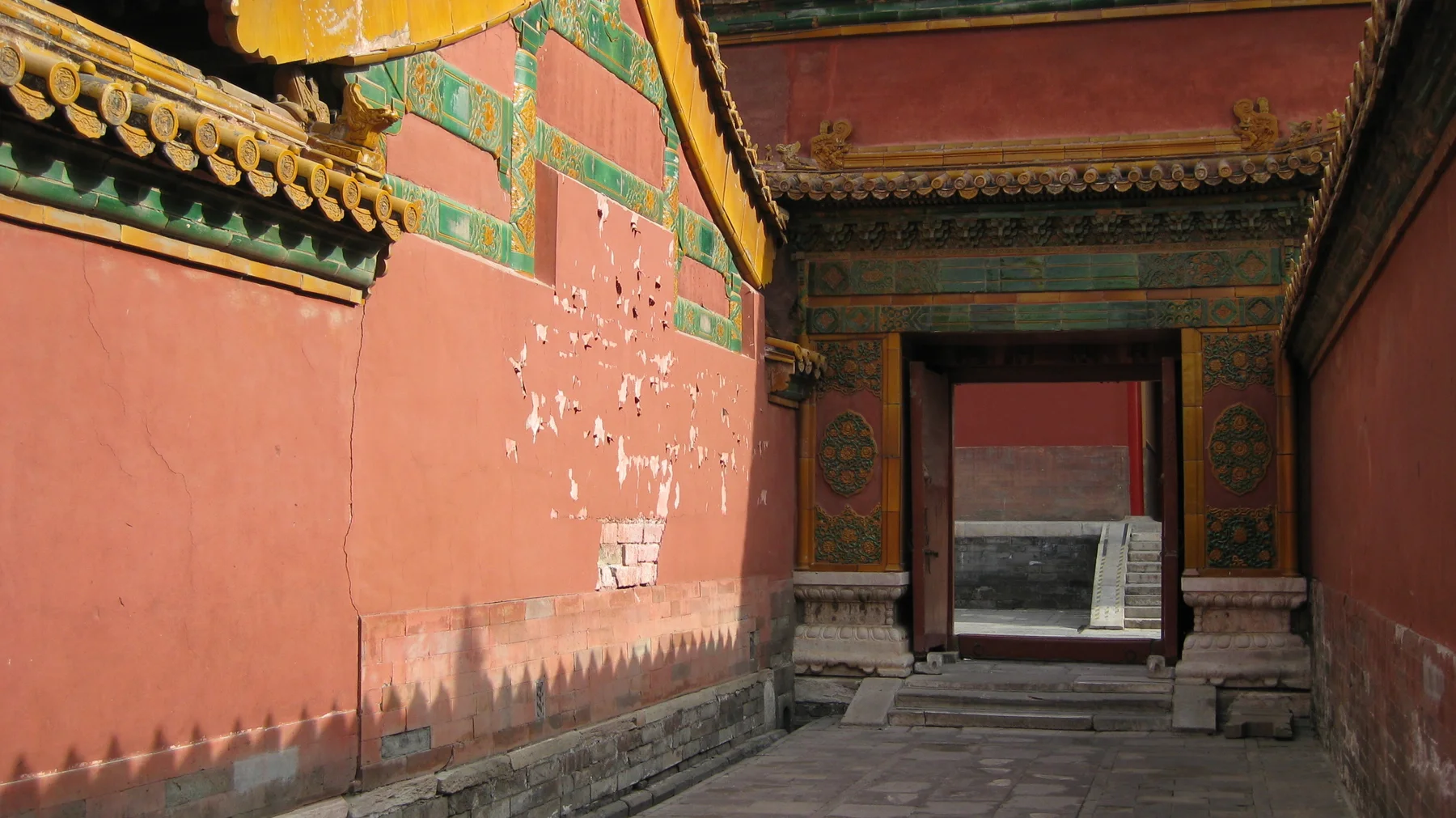 A paved passage runs beside a weathered red wall towards a rectangular doorway, with sunlight and pointed shadows across the wall.