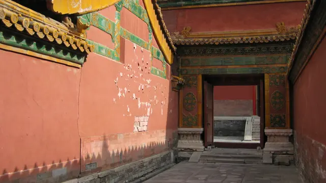 A paved passage runs beside a weathered red wall towards a rectangular doorway, with sunlight and pointed shadows across the wall.