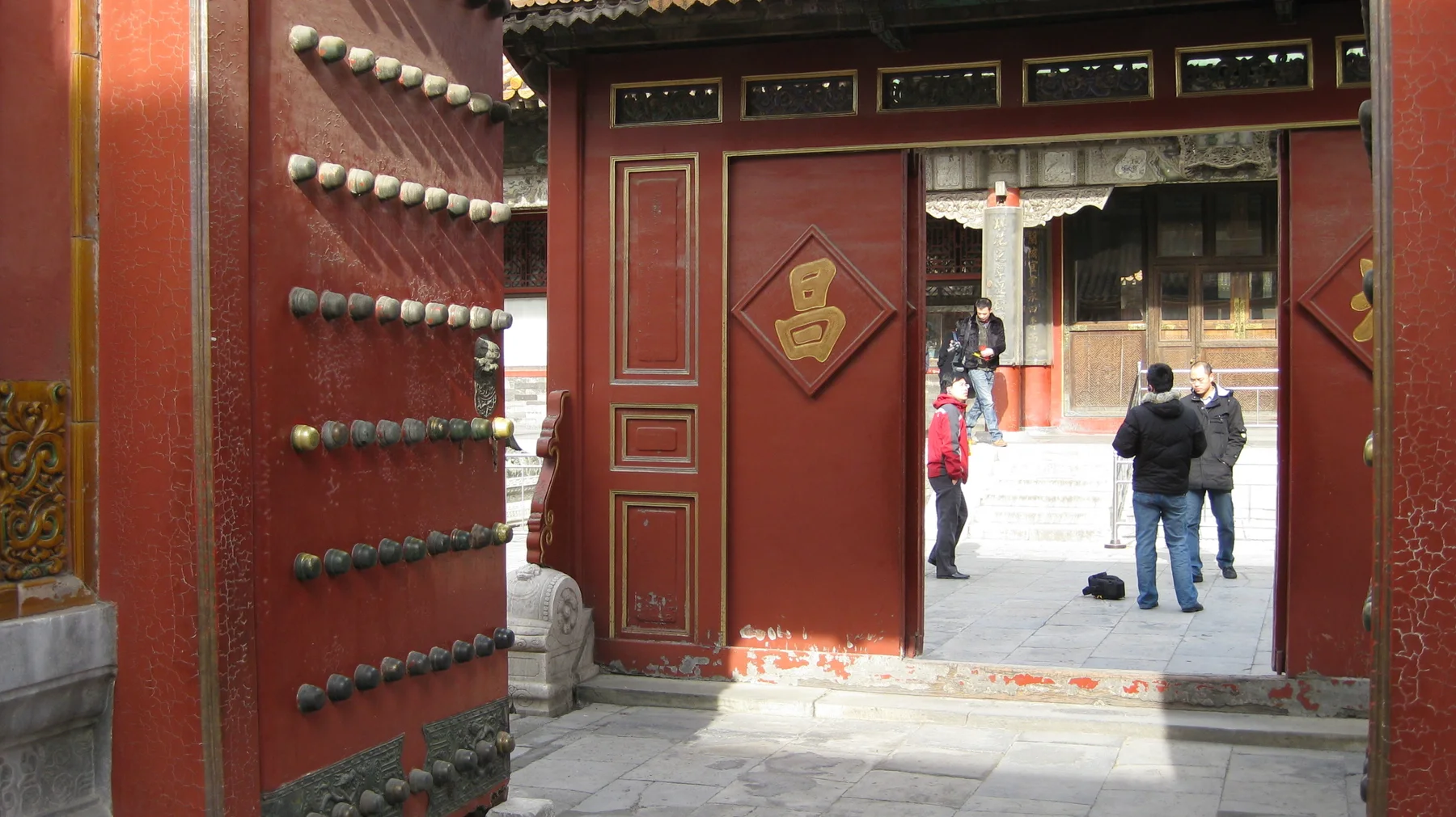 A heavy red door studded with metal knobs opens towards another doorway and a courtyard where two visitors stand.