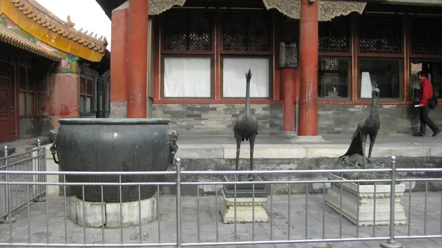 A large dark metal vessel and two tall bird sculptures stand behind railings in front of a building with red columns.