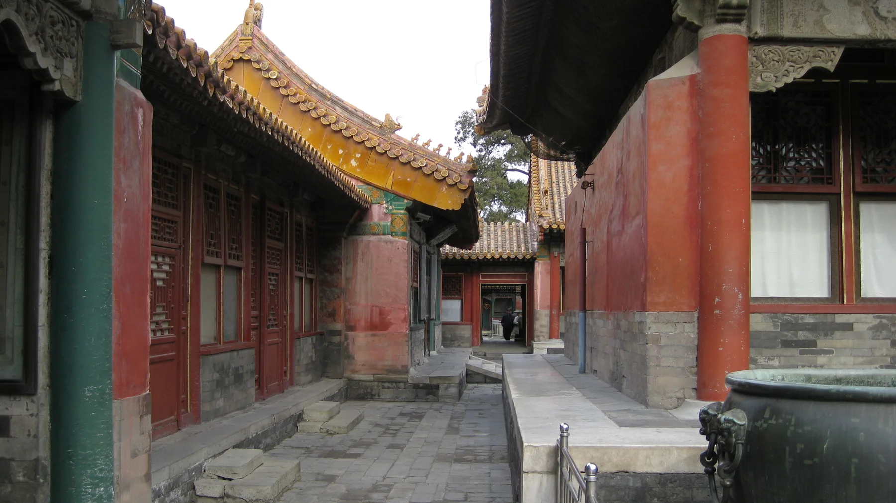A narrow paved passage lies between red-columned buildings with tiled roofs, with part of a dark metal vessel in the foreground.