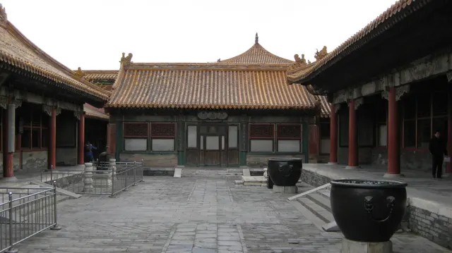 Large dark vessels stand in a paved courtyard surrounded by red-columned buildings with golden tiled roofs.