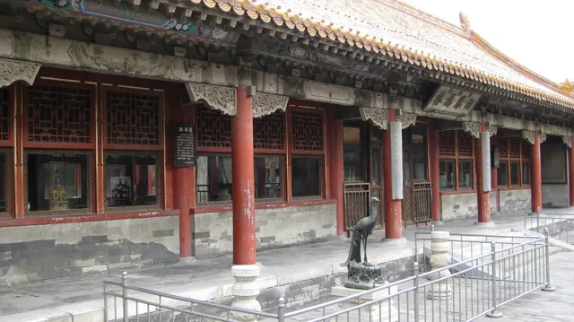 A long row of red columns supports decorated eaves above lattice windows, with a bird sculpture and railings in the courtyard.
