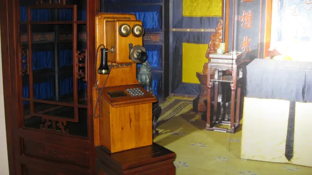 An old wooden telephone with two bells and a hanging receiver is displayed behind glass, with a room backdrop beyond.