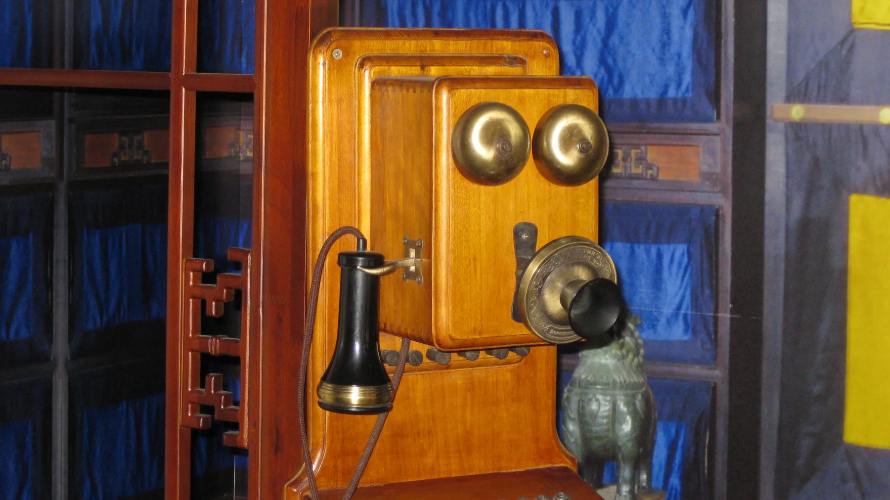 A close-up of a wooden telephone shows two round metal bells, a circular mouthpiece and a dark hanging receiver.