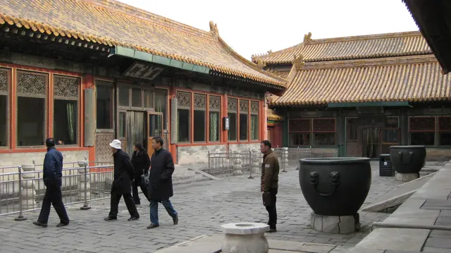 Visitors walk across a paved courtyard between red-columned buildings, with large dark vessels beside the path.