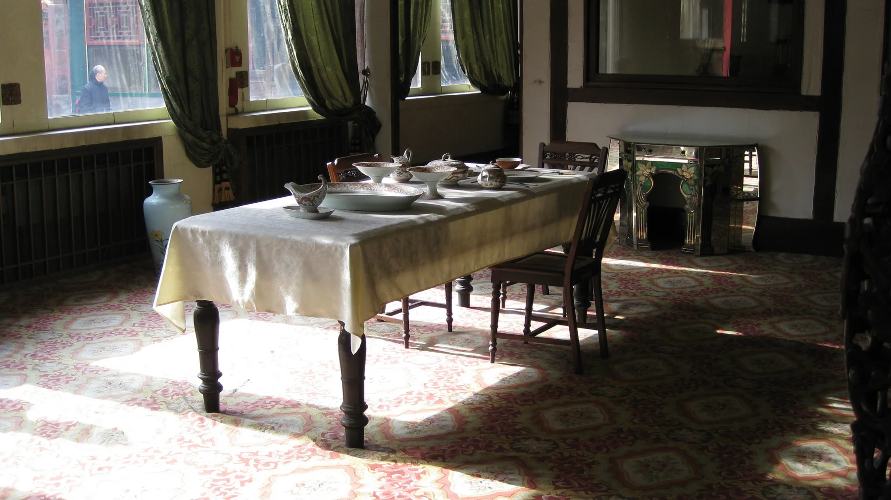 A cloth-covered dining table set with dishes stands beside tall curtained windows, with dark wooden chairs and sunlight on the patterned floor.
