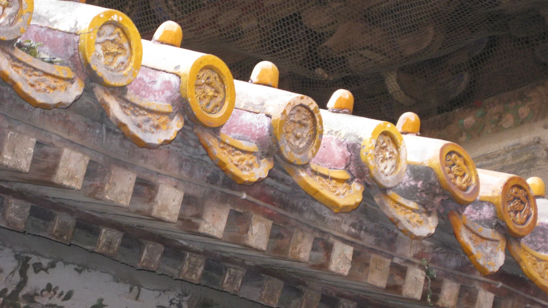 Round golden tile ends with raised patterns line the edge of a weathered roof above dark wooden brackets.