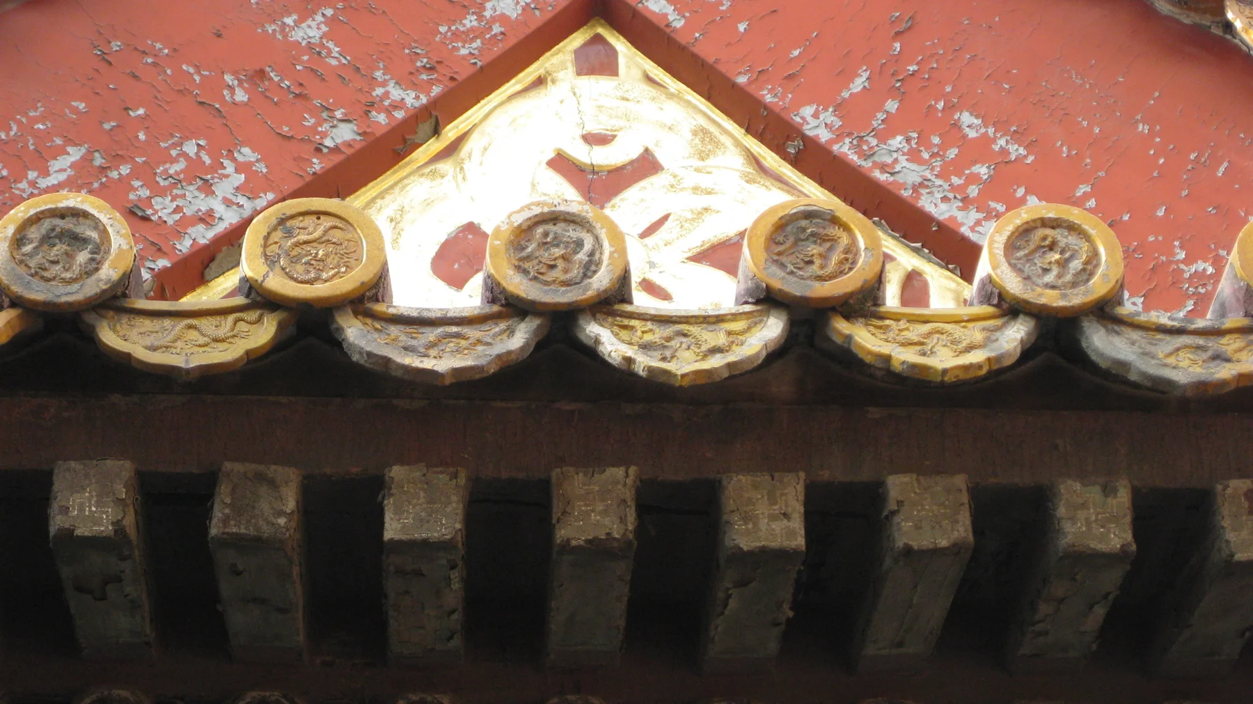 Golden tile ends and a triangular decorative panel stand out against peeling red paint above a row of dark roof supports.