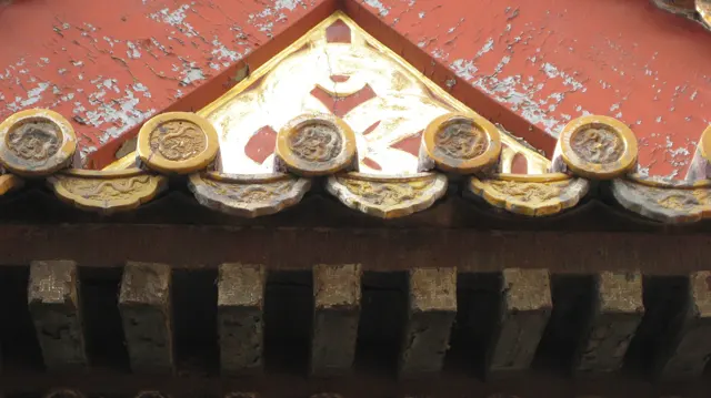 Golden tile ends and a triangular decorative panel stand out against peeling red paint above a row of dark roof supports.