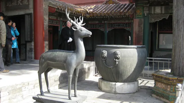 A dark deer sculpture stands beside a large metal vessel in a courtyard, with red columns and visitors behind.
