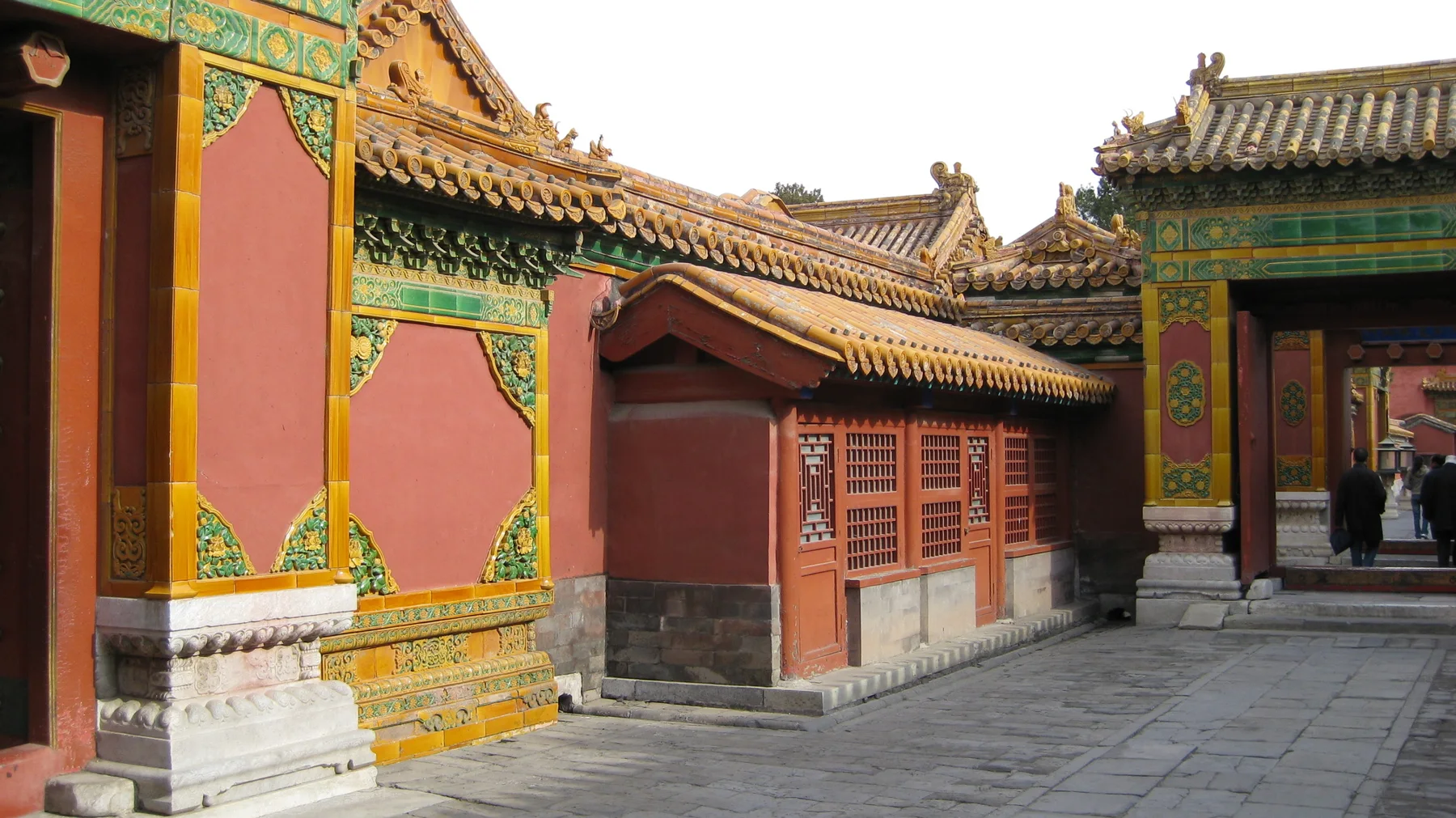 A paved passage runs between red walls and a small lattice-fronted structure, with ornate green-and-gold gateways at either end.