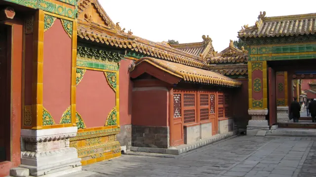 A paved passage runs between red walls and a small lattice-fronted structure, with ornate green-and-gold gateways at either end.