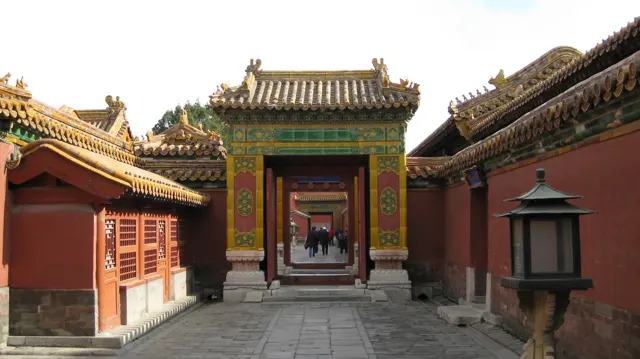 A green-and-gold gateway frames a series of doorways along a paved passage, with red walls and a dark lantern nearby.