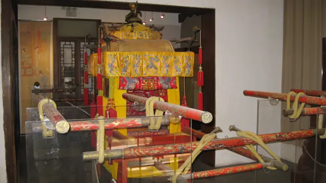 A red-and-yellow covered carrying chair is displayed behind glass, with long poles, ropes and decorative panels.