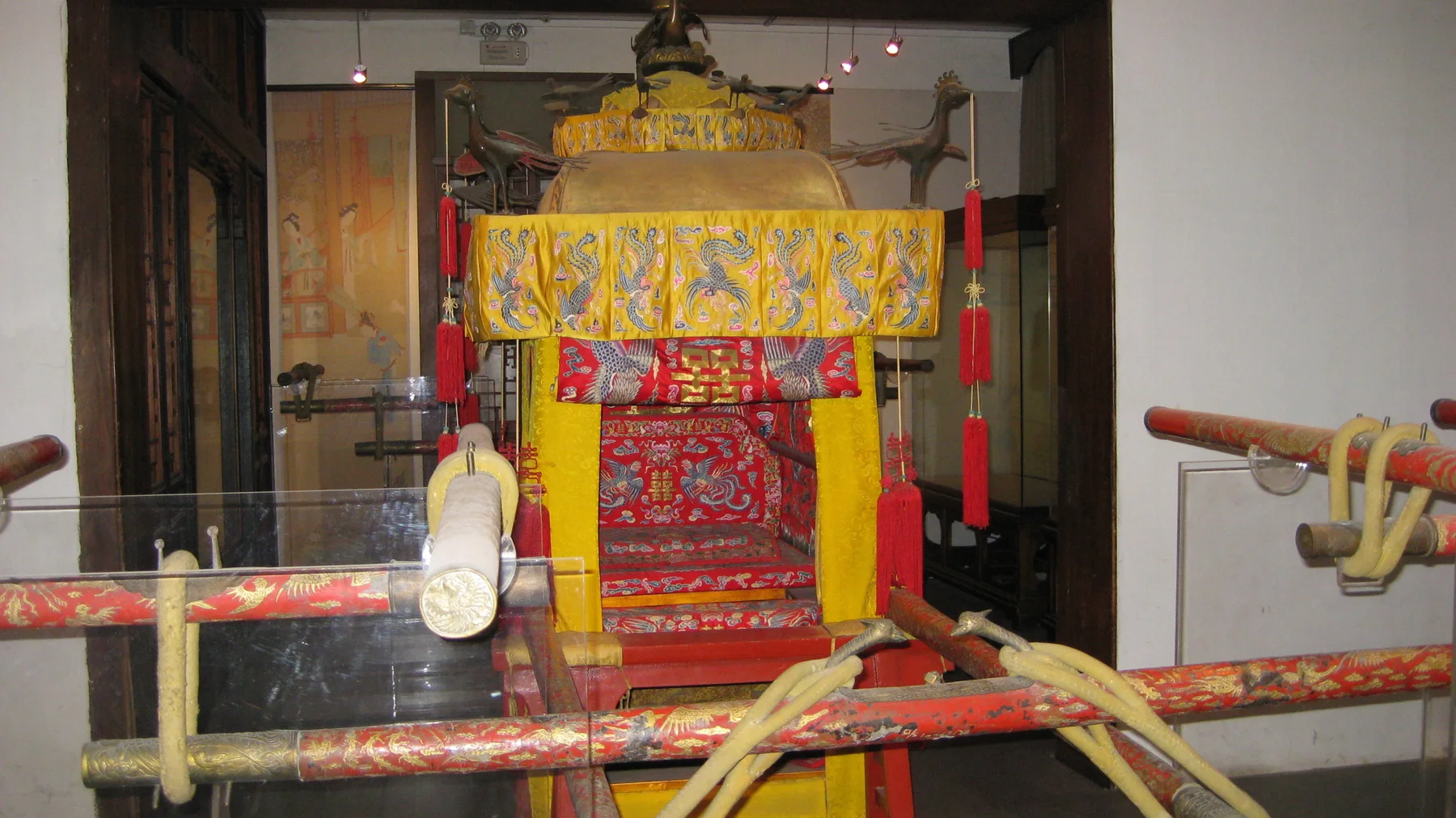 A closer front view of a covered carrying chair shows a yellow canopy, red patterned panels and long poles crossing the foreground.