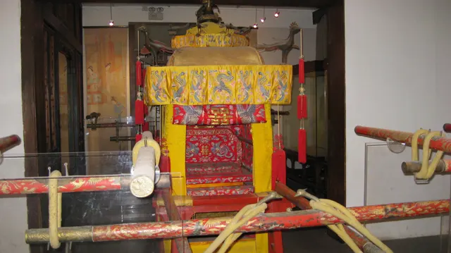 A closer front view of a covered carrying chair shows a yellow canopy, red patterned panels and long poles crossing the foreground.