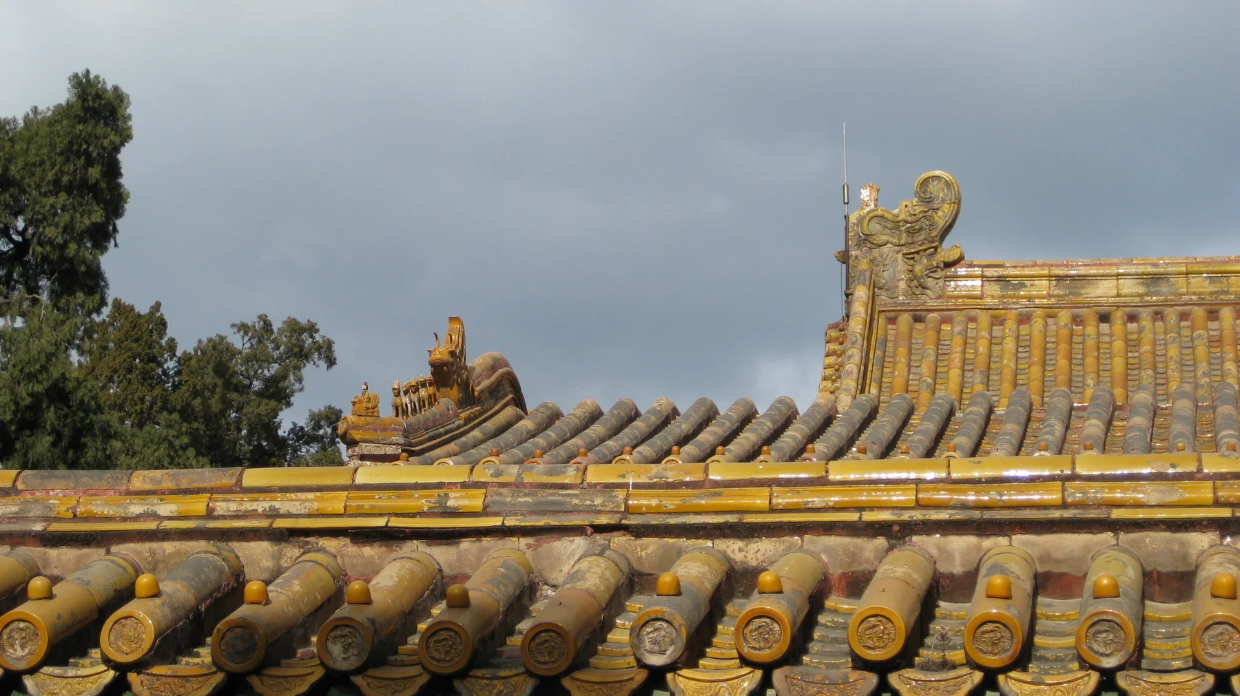 Golden roof tiles and decorative ridge figures stand against grey clouds, with green trees to the left.