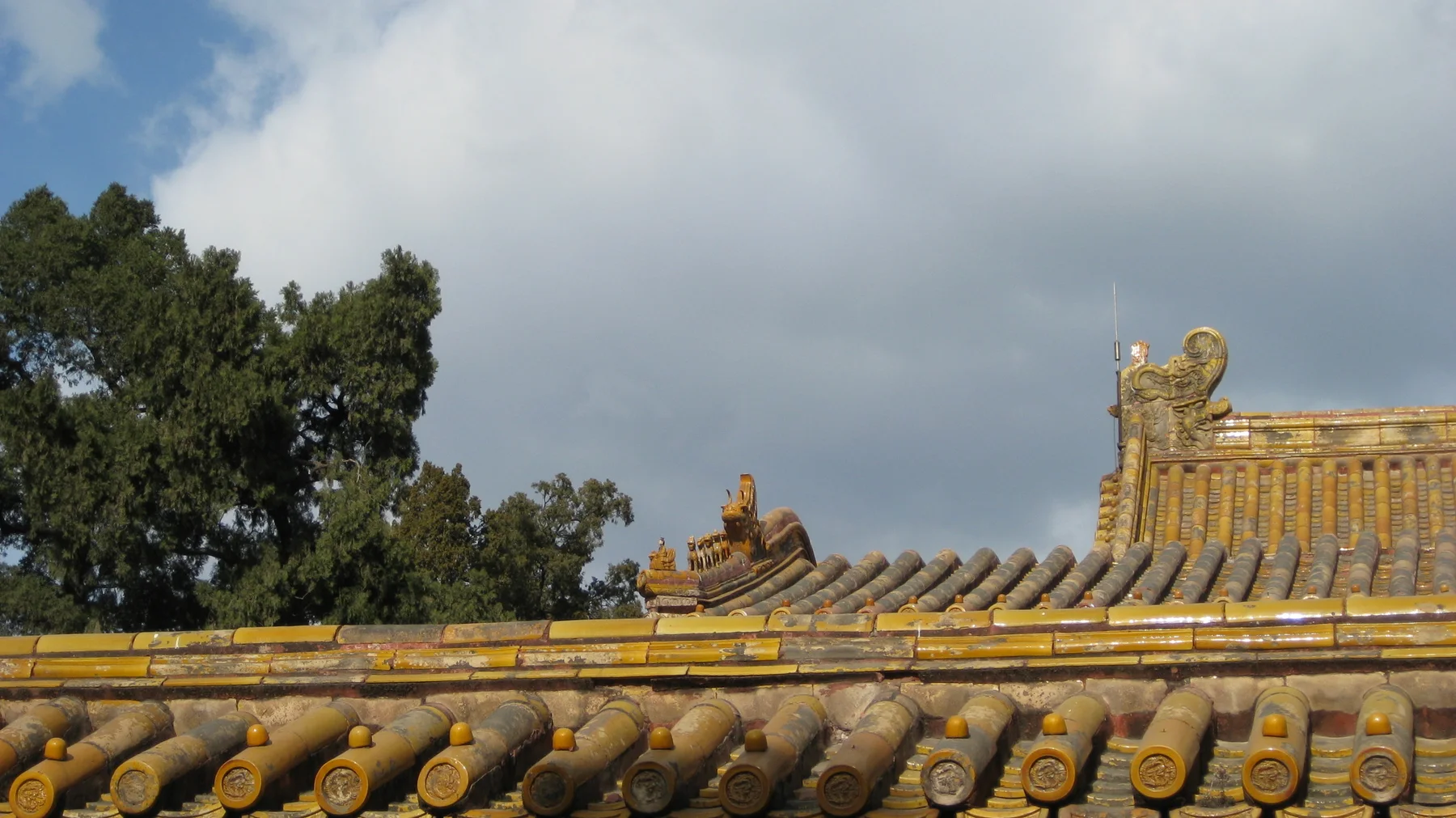 Rows of golden tiles and curled roof ornaments stretch beneath clouds and a small patch of blue sky, with trees beyond.