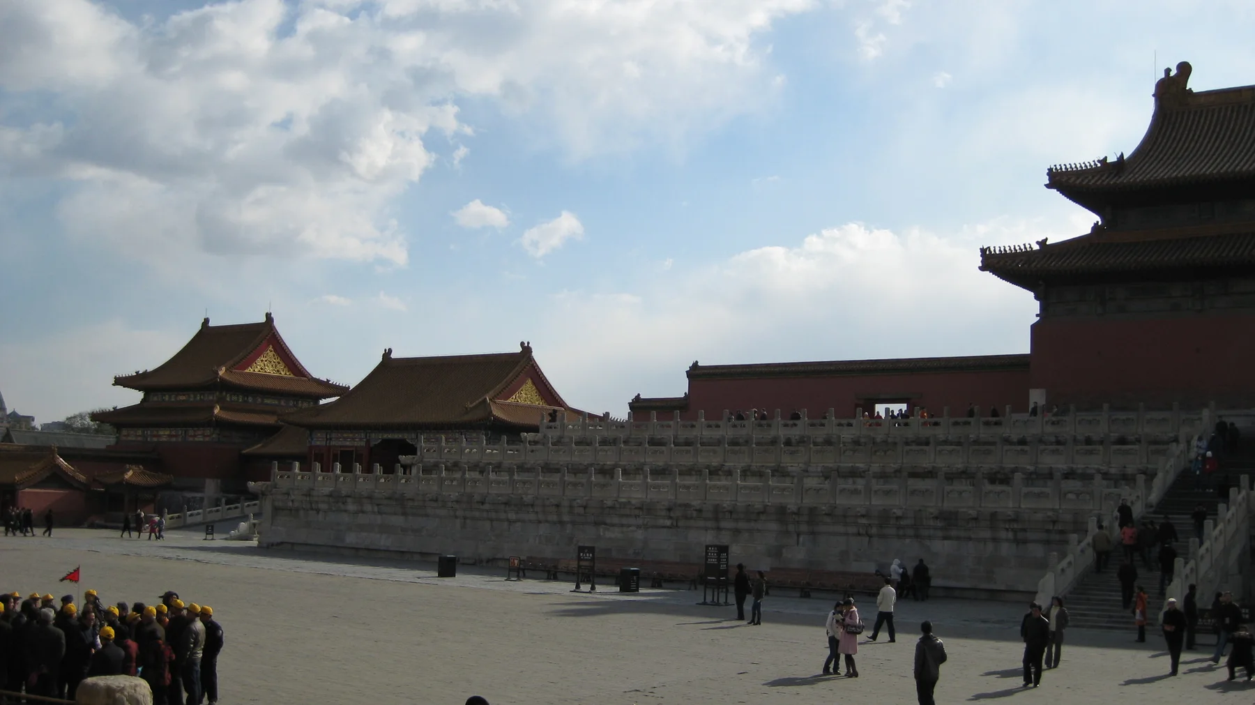 Visitors cross a wide courtyard beneath layered stone terraces and palace rooflines, with part of a large hall in shadow at the right.