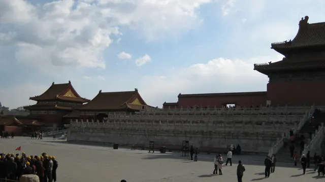 Visitors cross a wide courtyard beneath layered stone terraces and palace rooflines, with part of a large hall in shadow at the right.