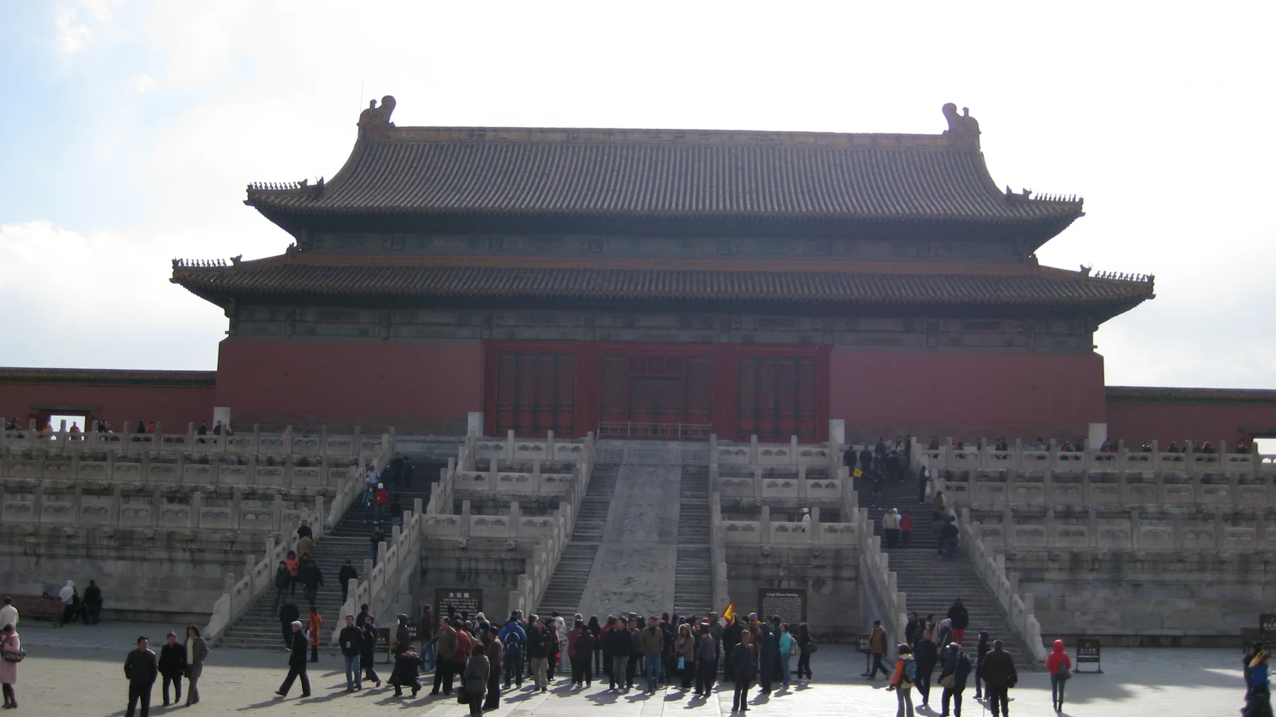 Visitors gather below broad stone stairways leading to a large red hall with a double-tiered tiled roof, backlit by a bright sky.