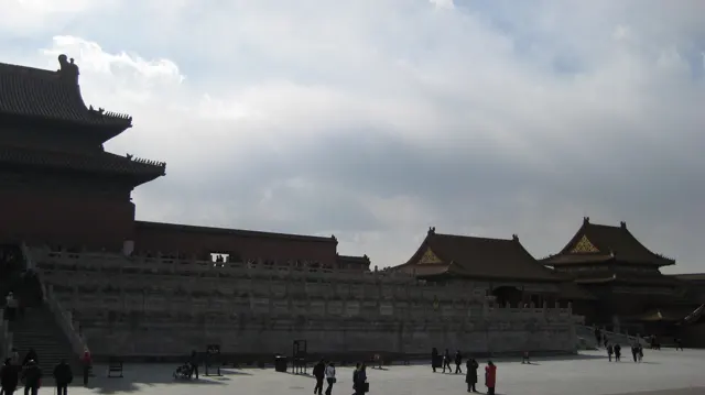 A long raised stone terrace runs beneath palace buildings, with visitors crossing the open courtyard below a cloudy sky.