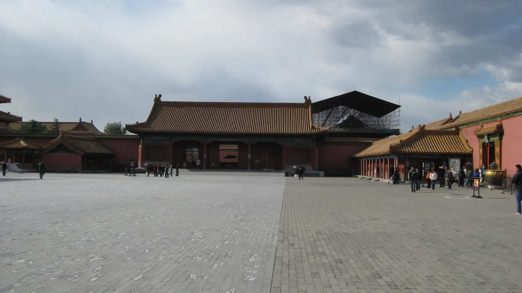 A broad paved courtyard extends towards red buildings with golden roofs, with a few visitors along the far edge beneath heavy clouds.
