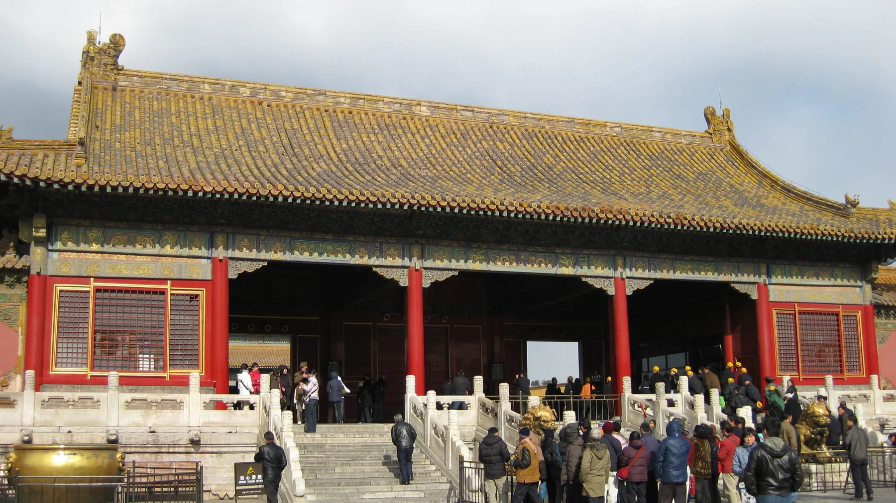 Visitors pass through a red-columned gateway beneath a golden roof, with carved pale stone railings bordering the steps.