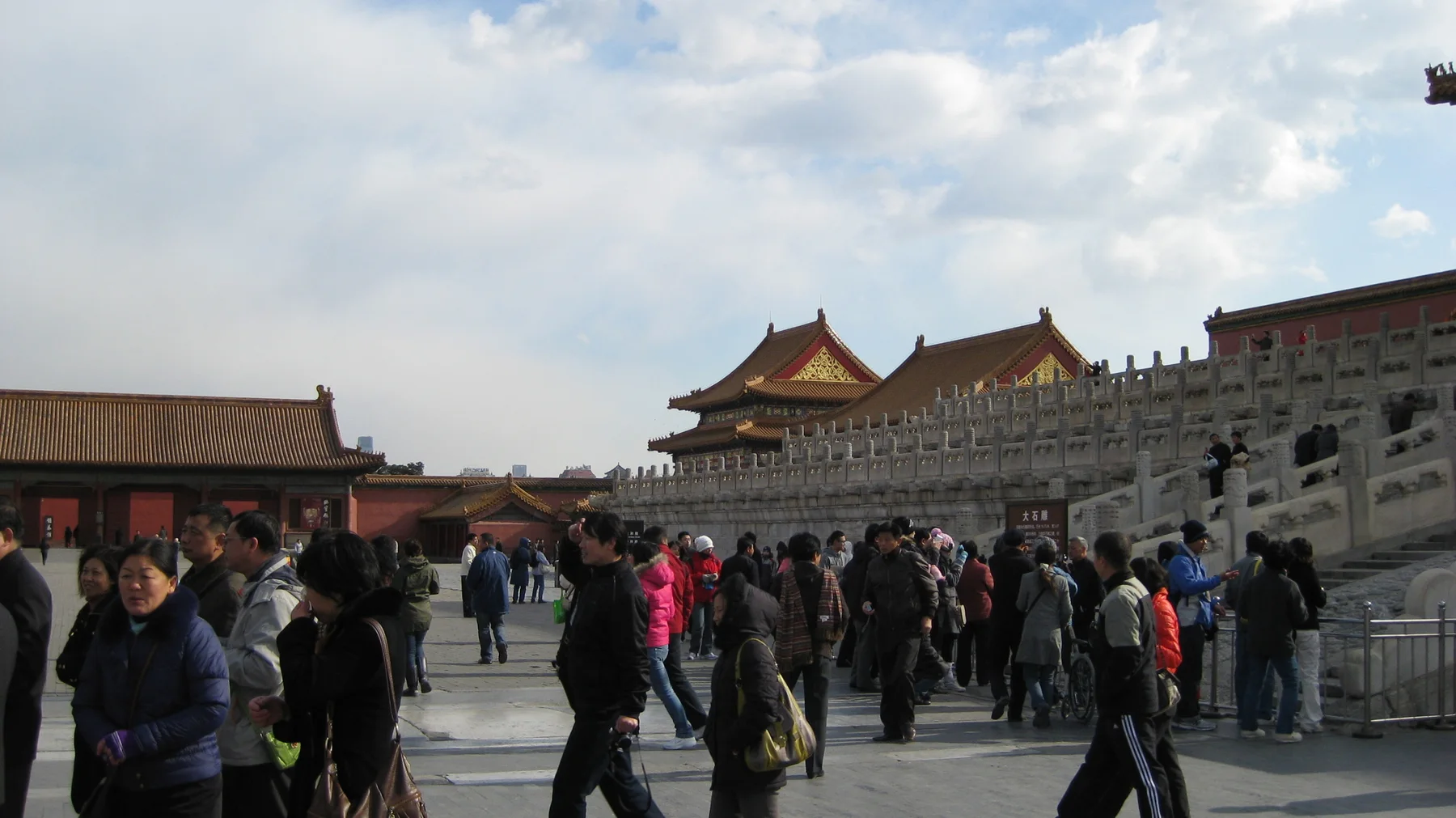 Groups of visitors walk and gather alongside a high stone terrace topped by balustrades and golden-roofed buildings.