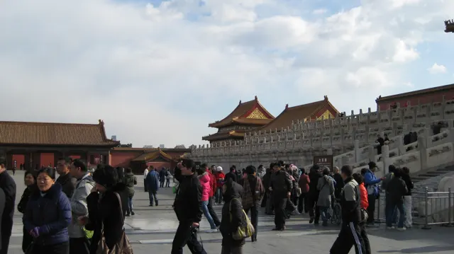 Groups of visitors walk and gather alongside a high stone terrace topped by balustrades and golden-roofed buildings.