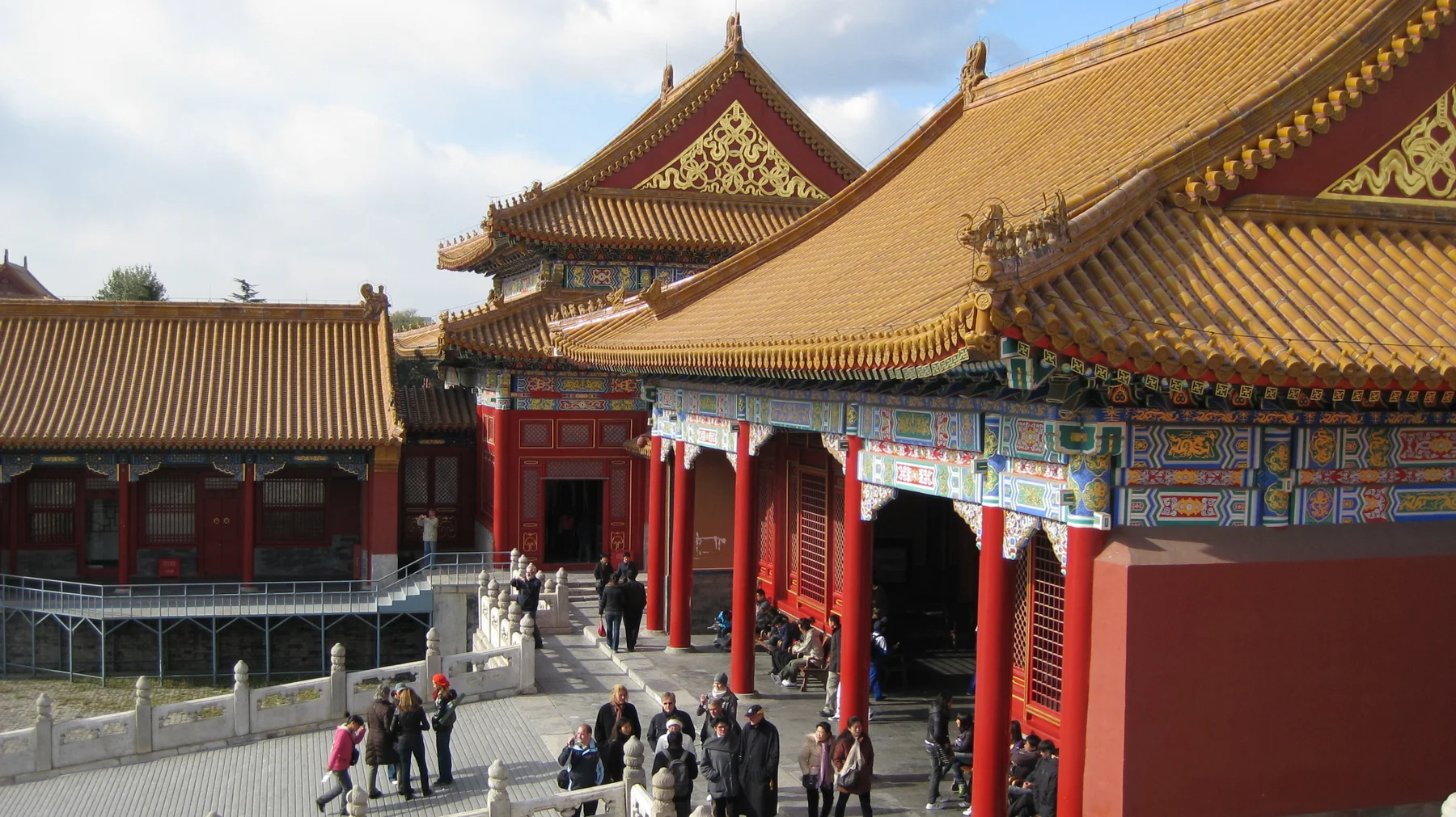 An elevated view follows a richly painted facade with red columns and layered golden roofs, with visitors moving below.