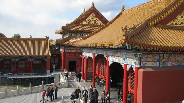 An elevated view follows a richly painted facade with red columns and layered golden roofs, with visitors moving below.