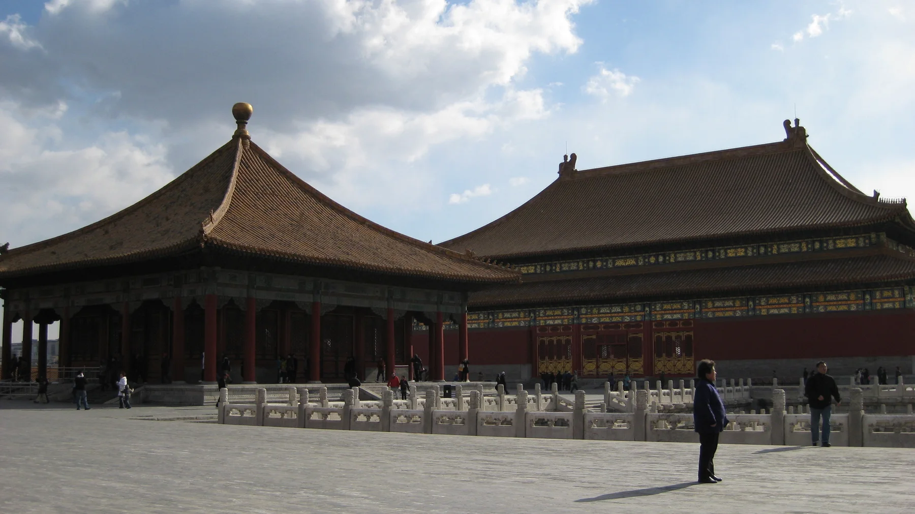 A pavilion with a pointed tiled roof stands beside a larger hall, separated from the open courtyard by pale stone balustrades.