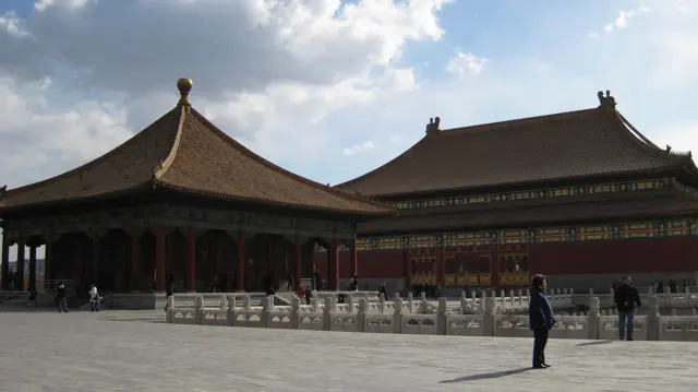 A pavilion with a pointed tiled roof stands beside a larger hall, separated from the open courtyard by pale stone balustrades.