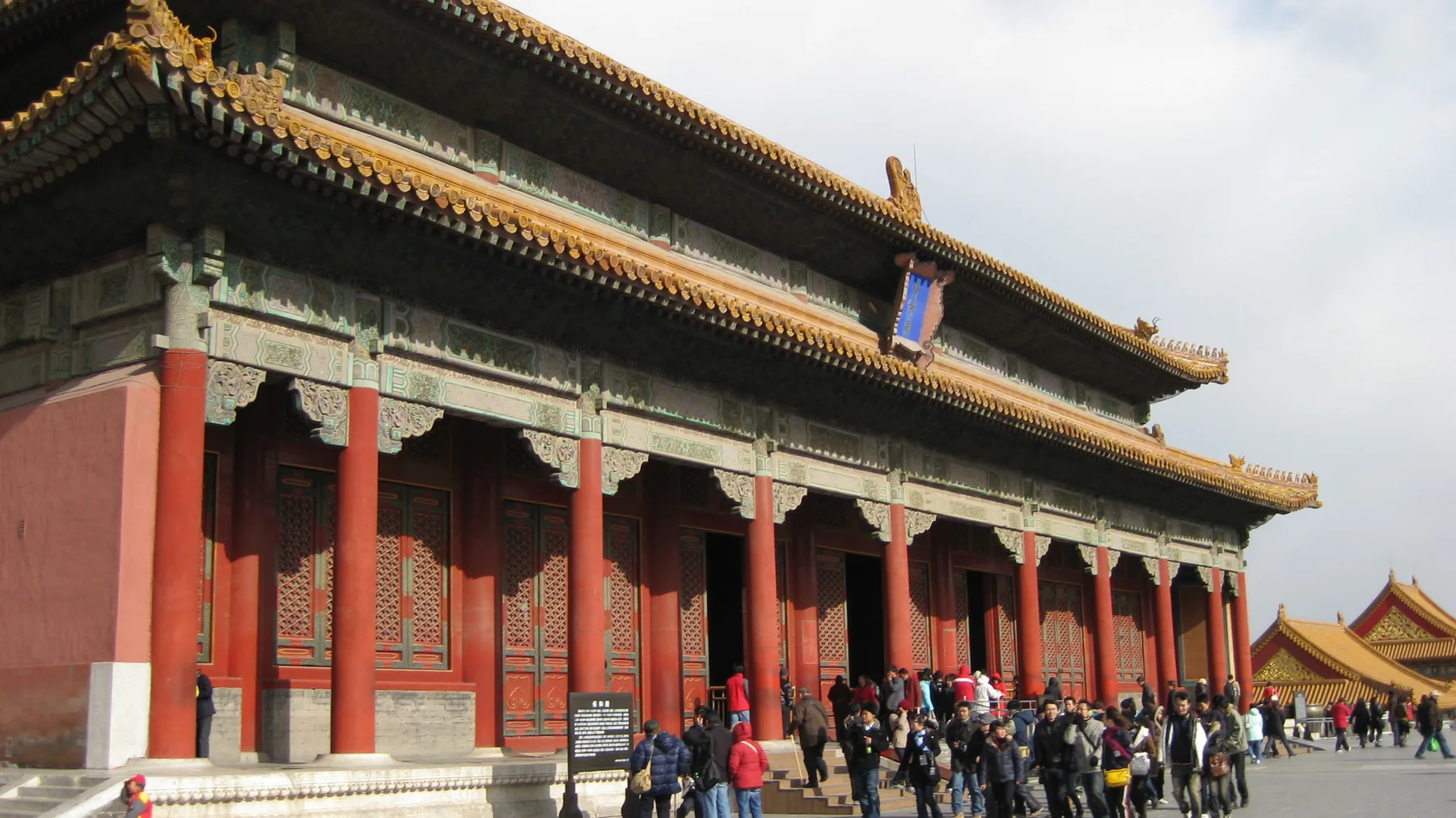 Visitors gather beside a long red-columned hall beneath decorated beams and a double-tiered golden roof.