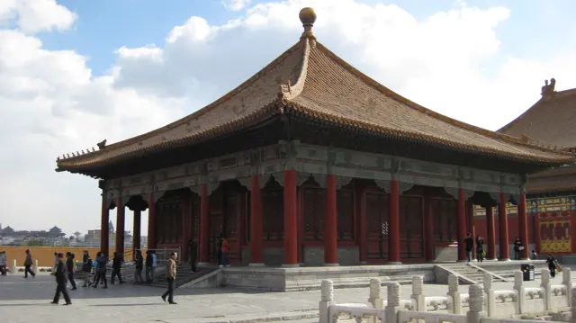 A square pavilion with red columns and a sweeping tiled roof stands in sunlight, with visitors and pale stone railings around it.