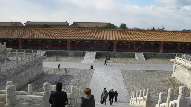 Visitors descend stone steps towards a wide paved courtyard, facing a long red gallery with a golden roof.