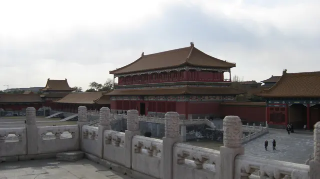 A carved stone balustrade frames a view towards a red building with a broad tiled roof across a lower courtyard.