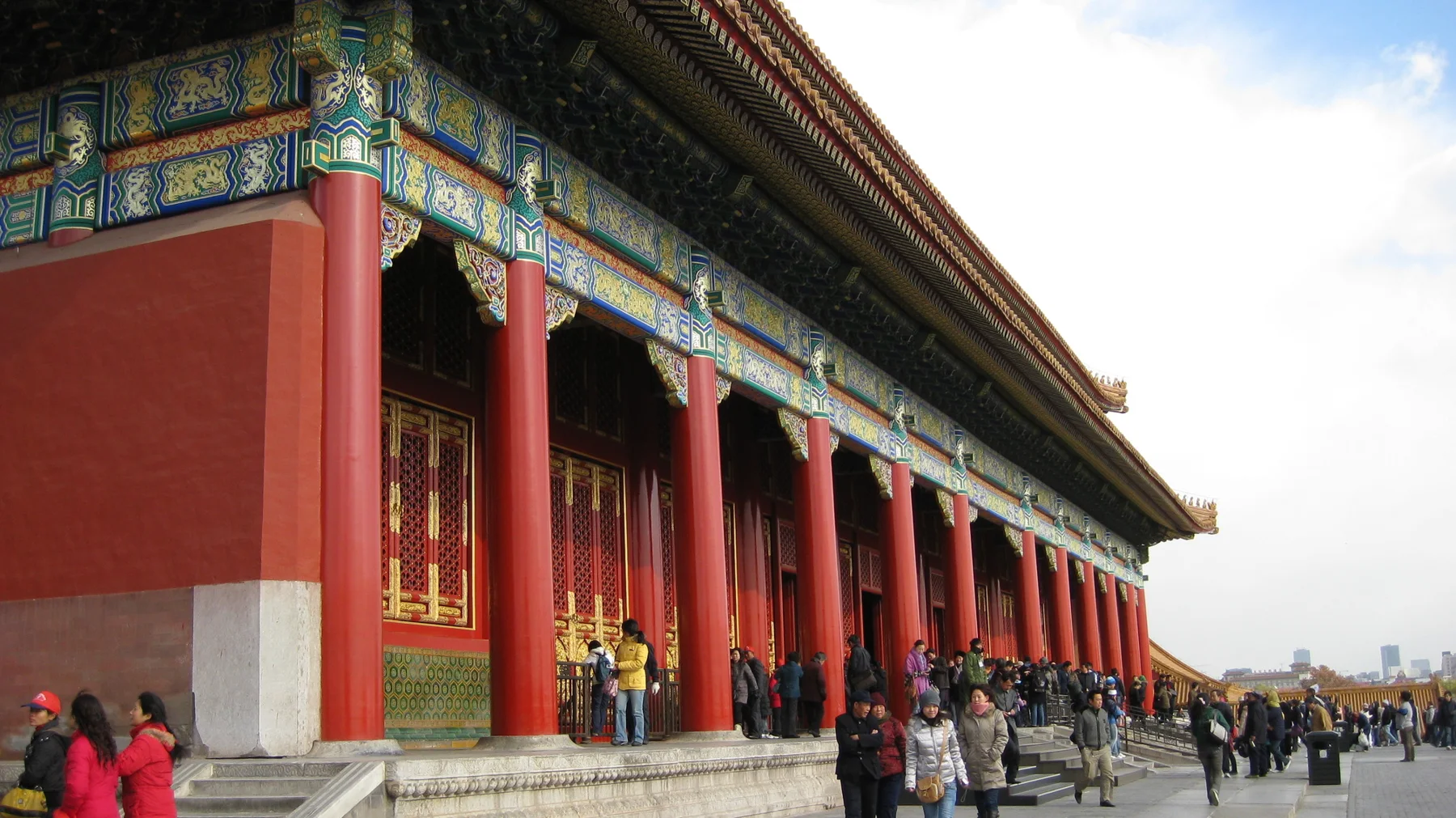A long row of red columns supports colourful painted beams, with visitors walking along the building and across the paving.