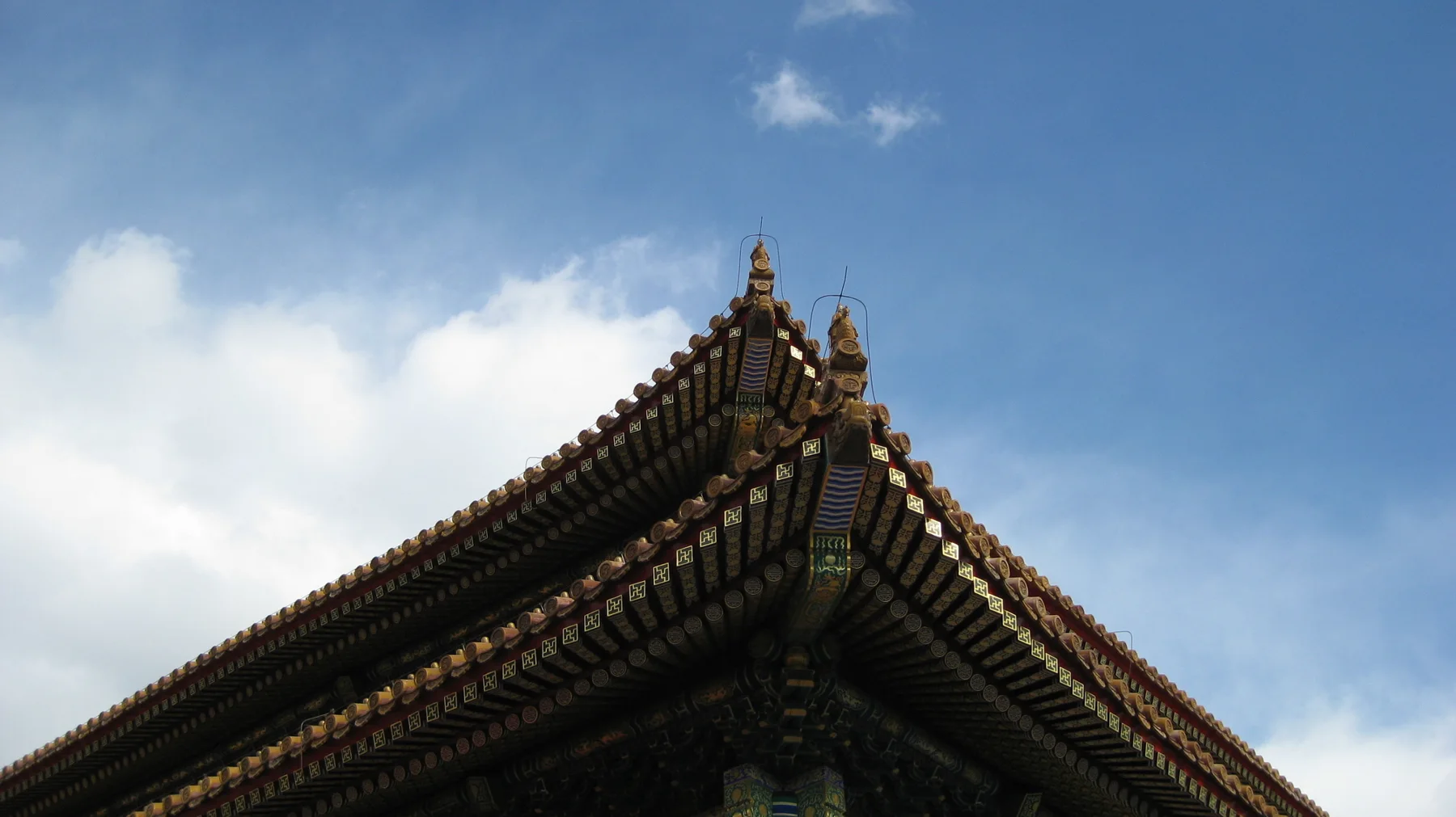 Two tiled roof slopes meet at a pointed corner with small ornaments, seen against a blue sky and white clouds.