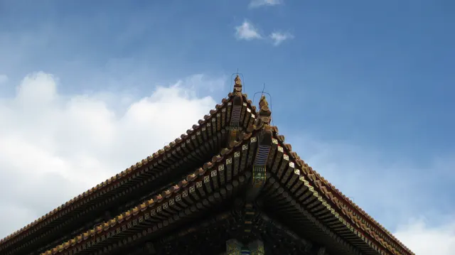 Two tiled roof slopes meet at a pointed corner with small ornaments, seen against a blue sky and white clouds.