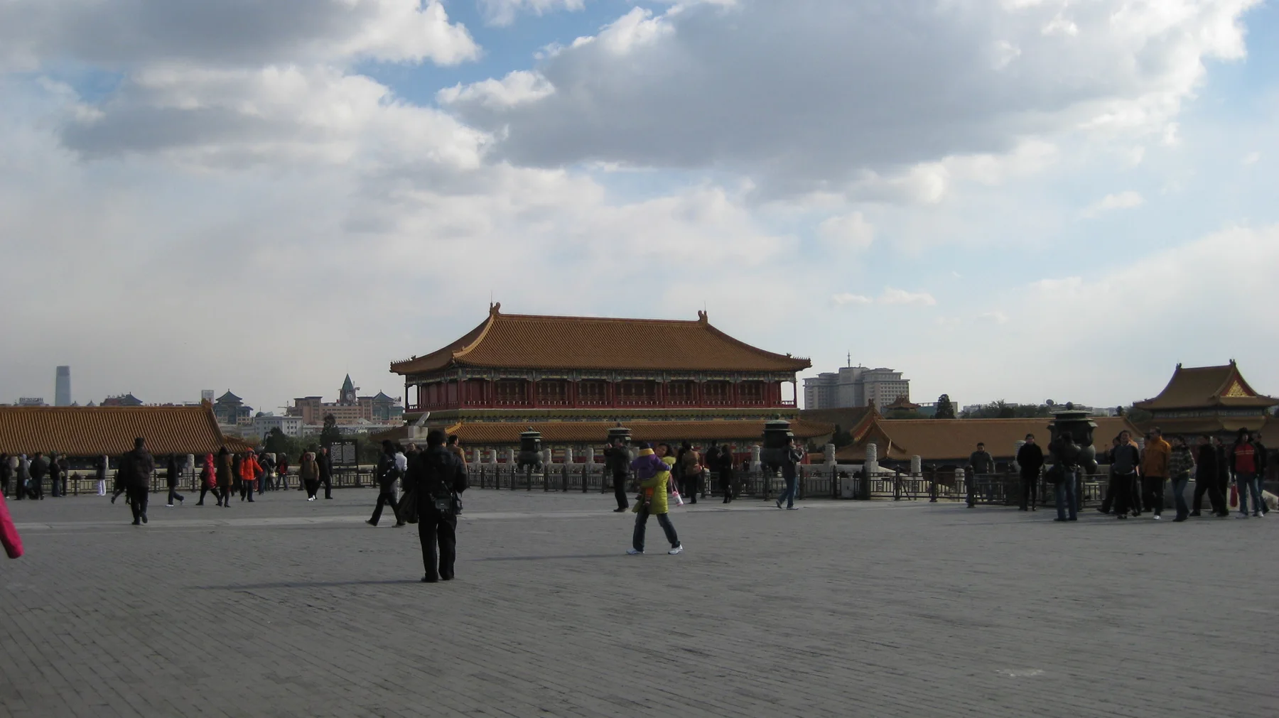 Visitors stand and walk across an expansive courtyard, with a distant golden-roofed building beneath sunlit clouds.