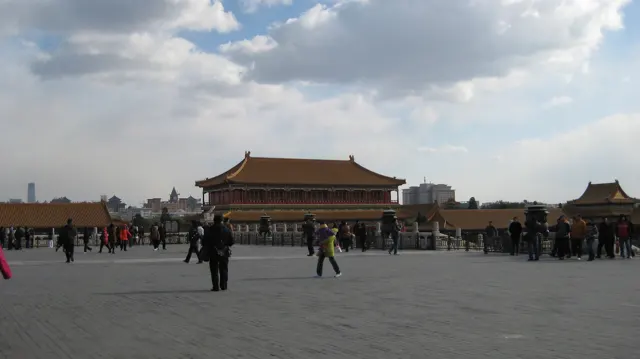 Visitors stand and walk across an expansive courtyard, with a distant golden-roofed building beneath sunlit clouds.