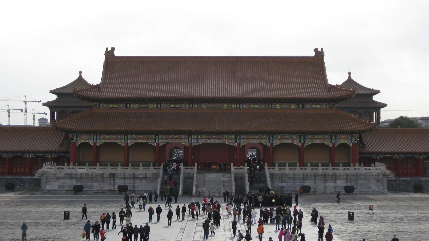 A large red gateway with layered tiled roofs faces visitors crossing the stone-paved space in front of it.